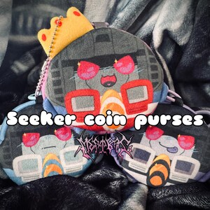 May include: Three plush coin purses featuring stylised robot character faces with red visor-like eyes, grey and blue details, and a yellow crown on the top one. The text Seeker coin purses is printed in white, and a logo is visible below it.