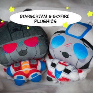 May include: Two plushies labelled STARSCREAM & SKYFIRE PLUSHIES. The left plush is dark grey with red accents and pink sunglasses. The right plush is white and grey with blue accents and blue sunglasses. Both feature small bodies and cartoonish expressions.