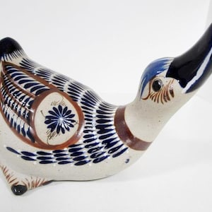 May include: A ceramic duck figurine with a cream-colored body, adorned with intricate blue and brown painted details. The duck features a long, upward-curving black beak, blue head, and a floral design on its side. The figurine is a decorative item.