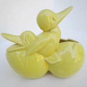 May include: A yellow ceramic planter shaped like two birds, one perched on top of the other. The planter has a smooth, glossy finish.