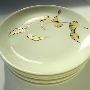 May include: A stack of six off-white ceramic plates. Each plate features a gold leaf and vine design. The leaves have detailed veining, and the vines are thin and elegant. The plates have a simple, classic design.