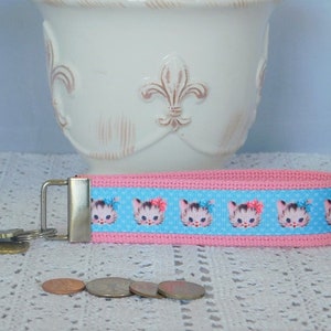 May include: Pink and blue key chain with a repeating pattern of cartoon kittens with pink bows. The key chain has a silver metal ring.