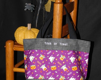 Op de afbeelding: Een grijze Halloween-draagtas met een paarse band met een patroon van geesten, pompoenen en snoepmaïs. De woorden "Trick or Treat" staan bovenaan gedrukt. De tas heeft zwarte handvatten.