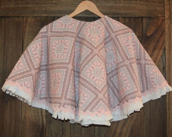 May include: A pink and white patterned cape with a scalloped edge and fringe trim. The cape is hanging on a wooden hanger.