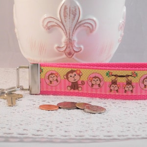 May include: Pink and yellow key chain with a repeating pattern of monkeys wearing flowers. The monkeys are brown and have pink cheeks. The key chain has a silver metal key ring.