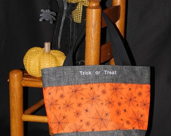 Op de afbeelding: Een Halloween-tote bag met een grijze boven- en onderkant en een oranje midden met zwarte spinnenwebben. De woorden "Trick or Treat" staan bovenaan gedrukt. Een houten kattenversiering en een pompoen staan op de achtergrond.