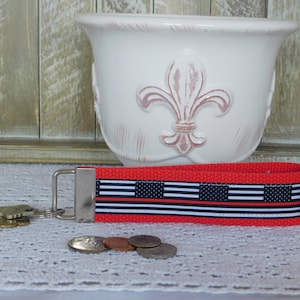 May include: Red and black key chain with a thin black and white ribbon with a thin red stripe. The ribbon has a pattern that resembles the American flag with a thin black line across the middle.