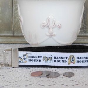 Puede incluir: Llavero negro y blanco con una cinta blanca con tres ilustraciones de perro de caza y el texto "Basset Hound".