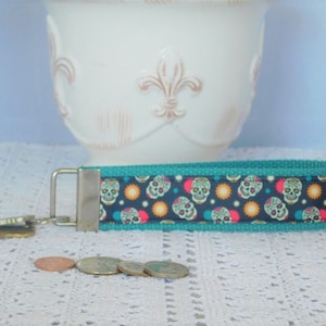 May include: Teal blue key chain with a silver key ring and a colorful fabric strap with a pattern of sugar skulls.