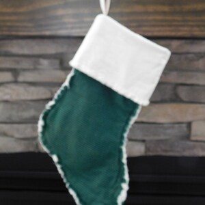 May include: A festive Christmas stocking, featuring a green body and a white cuff. The stocking is trimmed with white faux fur and hangs from a decorative branch. The stocking is made of a textured fabric.