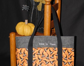 Op de afbeelding: Een grijze Halloween-draagtas met zwarte handvatten en een oranje paneel met zwarte heksenhoeden. De tekst "Trick or Treat" is gedrukt op de grijze stof. Een houten kat en pompoen staan op de achtergrond.