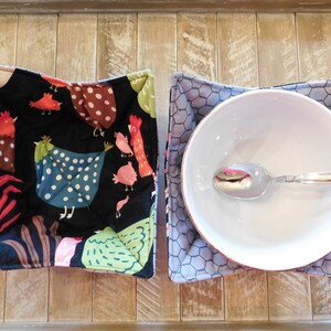 May include: Two fabric bowl cozies. One is black with a white polka dot pattern and features a colorful chicken design. The other is gray with a honeycomb pattern. A white bowl with a silver spoon is inside the gray cozy.