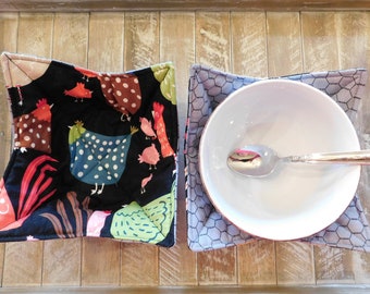 May include: Two fabric bowl cozies. One is black with a white polka dot pattern and features a colorful chicken design. The other is gray with a honeycomb pattern. A white bowl with a silver spoon is inside the gray cozy.