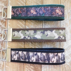 May include: Three keychains with different camouflage patterns. The top keychain has a green background with a brown and tan camouflage pattern. The middle keychain has a brown background with a green and tan camouflage pattern. The bottom keychain has a black background with a gray and tan camouflage pattern.