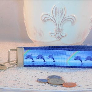 Puede incluir: Llavero azul y blanco con un diseño de delfines saltando sobre un arcoíris. El llavero está hecho de tela y tiene un anillo de metal plateado.