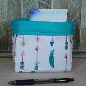 May include: A teal fabric storage bin with a pink and white arrow print. The bin has a teal fabric top and is sitting on a wooden surface. A pen is in front of the bin.