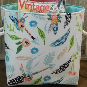 May include: A white canvas storage bin with rope handles. The bin has a teal blue lining and features a colorful pattern of feathers, arrows, and flowers. The bin is filled with a magazine titled "Vintage Style".