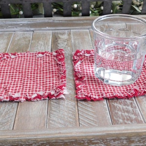May include: Two red and white gingham fabric coasters with frayed edges. A clear glass with water is sitting on one of the coasters.