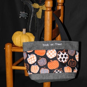 May include: A Halloween tote bag featuring a pumpkin pattern in orange, white, and black on a black background. The top edge reads "Trick or Treat". The bag is displayed on a wooden chair, next to a black cat decoration.