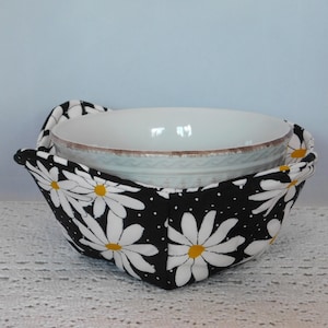 May include: A white ceramic bowl sits inside a black bowl cozy. The cozy is decorated with white daisies with yellow centers on a black background. The bowl cozy has two handles.