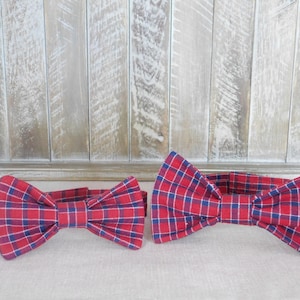 May include: Two red and blue plaid bow ties. The larger bow tie is on the right and the smaller bow tie is on the left.
