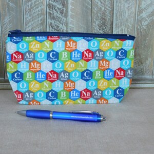 May include: A blue zippered pouch with a hexagonal pattern featuring the chemical symbols and names of elements. The pouch is on a light brown surface with a blue pen.