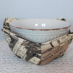 May include: A white ceramic bowl with a decorative rim sits inside a fabric bowl cozy. The cozy is beige and brown with a vintage newspaper print design. The cozy is designed to hold the bowl.