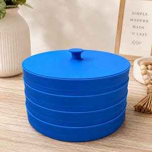 May include: A bright blue, circular, stackable container with a matching lid featuring a central knob handle. The container consists of three distinct, interlocking horizontal sections that form a cylindrical shape with smooth, solid-colored walls.