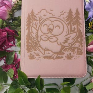 Puede incluir: Una funda rectangular de color rosa claro para tableta o libro con una ilustración grabada con láser de una criatura redonda similar a un pájaro con las alas extendidas, en un entorno forestal con pinos, arbustos y una seta.
