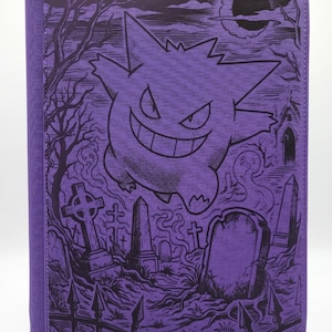 Peut inclure: Un classeur violet avec une illustration graphique noire représentant un personnage Gengar flottant au-dessus d'un cimetière avec des pierres tombales, une clôture, des arbres dénudés et un croissant de lune dans le ciel nocturne.