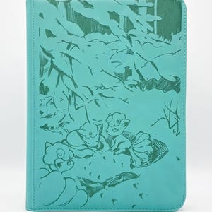 Peut inclure: Un classeur rectangulaire bleu sarcelle orné d'une illustration au trait vert foncé représentant trois personnages Vulpix se reposant sous des branches d'arbre stylisées. La couverture présente une surface texturée et des coins arrondis.