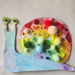 Op de afbeelding: Een slak-knutselwerk van papier met een lichtblauw lichaam met paarse stippen. Het huisje is een cirkelvormige collage van regenboogkleurig papier, versierd met diverse texturen zoals pluizige pompons, een gele knoop en kleine kraaltjes.
