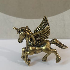 Puede incluir: Una pequeña figura de metal en tono latón de un unicornio alado en posición de galope. La criatura presenta un cuerno único en la frente, alas emplumadas detalladas extendidas hacia arriba y una cola fluida, con un acabado de estilo antiguo.