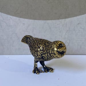 Pode incluir: Uma pequena estatueta metálica de cor dourada representando um pássaro com uma superfície texturizada semelhante a penas. O pássaro é retratado numa postura inclinada para a frente com a cabeça para baixo, exibindo padrões de plumagem detalhados e patas com garras.