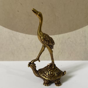 May include: A small brass figurine depicting a crane standing on top of a turtle. The crane has an elongated neck and textured wings, while the turtle features a detailed shell with geometric patterns. Both animals are rendered in a metallic gold tone.