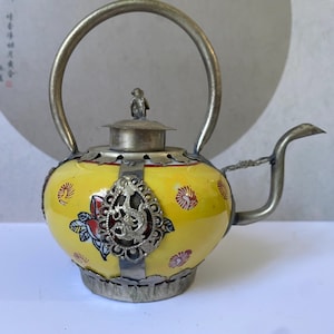 May include: A yellow ceramic teapot featuring a silver-toned metal handle, spout, and base. The body is decorated with red and blue floral patterns and a central silver-toned filigree overlay depicting a dragon. A small metal finial sits on the lid.