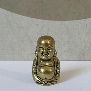 May include: A small, metallic gold-colored figurine of a laughing Buddha with a round belly, smiling face, and bald head. The figure is depicted in a seated position with hands resting on the lap, featuring detailed robes and a smooth, polished finish.