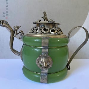 May include: A green jade teapot featuring ornate silver-toned metal accents, including a dragon-shaped spout, a lion head relief on the body, and a lid topped with a small monkey figure. The metal frame secures the jade segments with intricate detailing.