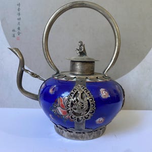 May include: A cobalt blue teapot featuring a silver-toned metal frame, a curved spout, and a high arched handle. The lid is topped with a small monkey figurine. The body is decorated with floral patterns and a central silver filigree dragon medallion.