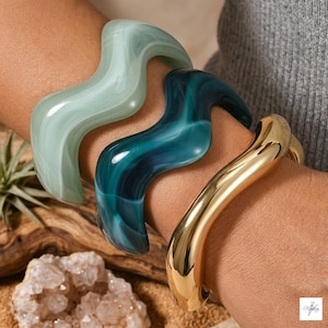 Pode incluir: Três pulseiras onduladas usadas juntas. Uma pulseira é verde-menta pálido com acabamento marmorizado, a segunda é verde-azulado profundo com acabamento marmorizado e a terceira é de metal dourado polido e reflexivo.