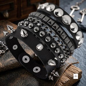 Könnte beinhalten: Ein Set aus schwarzen Armbändern mit verschiedenen metallischen Verzierungen, darunter silberfarbene konische Nieten, pyramidenförmige Nieten und ein dunkles Metallkettenband. Die Armbänder bestehen aus schwarzem Material und werden mit Druckknöpfen geschlossen.