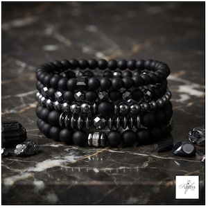 Könnte beinhalten: Ein Stapel aus fünf Perlenarmbändern mit mattschwarzen runden Perlen, facettierten dunkelmetallischen Perlen und glatten gunmetalfarbenen Zwischenelementen. Ein Armband enthält eine silberfarbene zylindrische Perle mit kleinen klaren Kristallen.