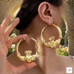 May include: A pair of large gold-toned hoop earrings featuring a Claddagh design. Each earring displays a central heart topped with a crown and flanked by two hands, integrated into the circular hoop structure with a polished metallic finish.