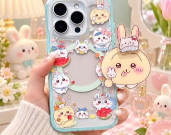 May include: A clear smartphone case with a light blue border featuring various cartoon animal stickers, including rabbits and cats, and a large circular magnetic ring. A prominent, round, beige cartoon rabbit character is attached to the side.