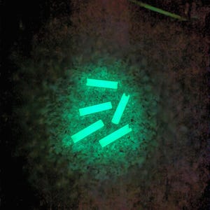 Peut inclure: Quatre petits bâtonnets rectangulaires en plastique phosphorescent émettent une lumière vert néon vive sur un fond sombre et texturé. Les bâtonnets sont disposés de manière éparse, chacun présentant une petite encoche à une extrémité.