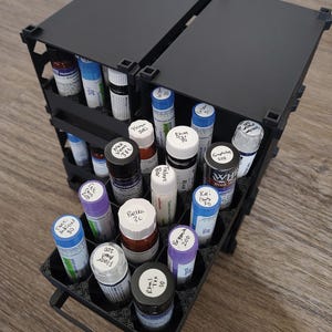 May include: A black modular storage organizer with multiple hexagonal compartments holding various small cylindrical glass vials. Each vial features a white cap and a label with handwritten text, organized in a tiered, open-front display rack.