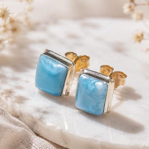 Puede incluir: Un par de pendientes de botón cuadrados con piedras azul claro pulidas engastadas en marcos de metal plateado con cierres de mariposa dorados. Las piedras presentan una textura lisa y veteada con diversos tonos de azul cielo y blanco.