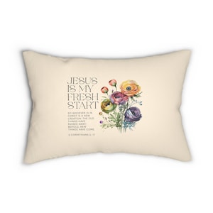 May include: A rectangular cream-coloured throw pillow featuring a watercolour floral bouquet in shades of yellow, purple, and green. The text reads: JESUS IS MY FRESH START. SO WHOEVER IS IN CHRIST IS A NEW CREATION. 2 CORINTHIANS 5:17.