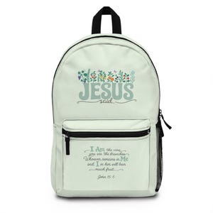 Puede incluir: Una mochila de color crema claro con un gráfico de ilustraciones botánicas coloridas sobre el texto "JESUS SAID..." y una sección inferior con el versículo "I Am the vine you are the branches Whoever remains in Me and I in him will bear much fruit John 15:5" en una fuente de color verde azulado oscuro.