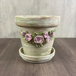 May include: A tapered plant pot with a matching saucer, featuring a distressed sage green finish with white highlights. The exterior is decorated with a central relief sculpture of light purple roses and green leaves arranged in a horizontal cluster.